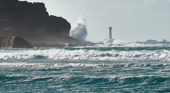 This landscape photograph captures the rugged Longships cliffs and powerful waves crashing against the coast at Sennen Cove in Cornwall during a windy autumn afternoon. In the background, the well-known Longships Lighthouse rises from the sea, standing as a sentinel amidst swirling spray and turbulent waters. The scene showcases the natural beauty of the United Kingdom’s coastline, with the dramatic interaction between the cliffs, the restless sea, and surrounding elements illustrating the untamed nature typical of Cornwall. The iconic lighthouse acts as a focal point, highlighting the maritime heritage of this region along the Atlantic coast.